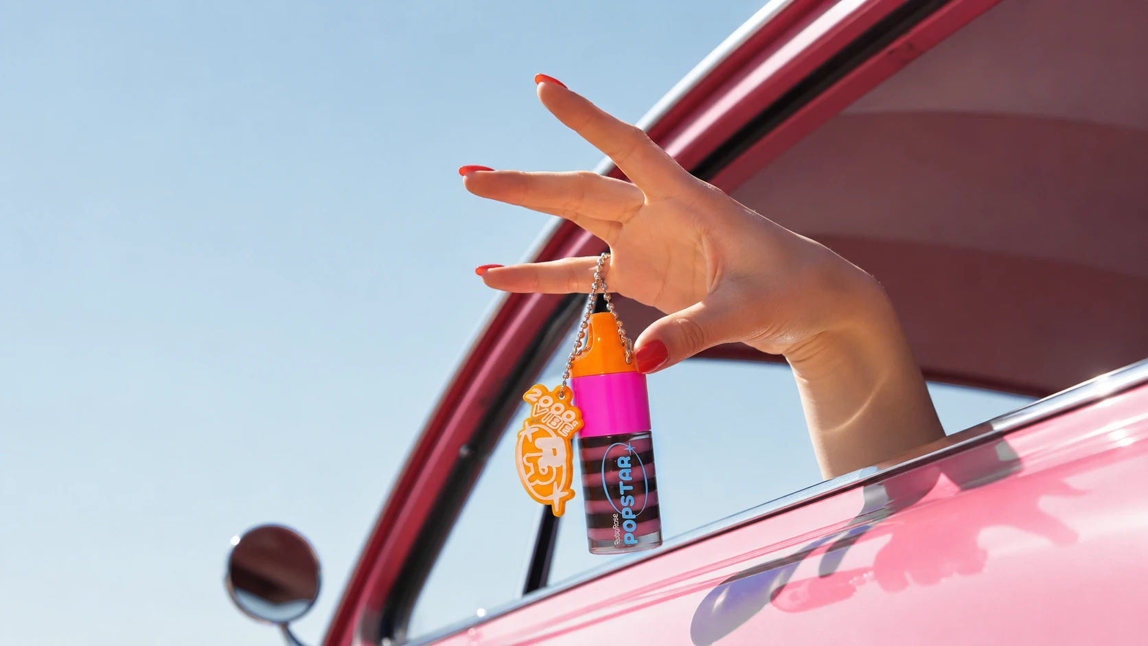 A hand out of a pink car window holding a Ruby Rose Popstar lip gloss on a 2000s Vibe keychain against a blue sky